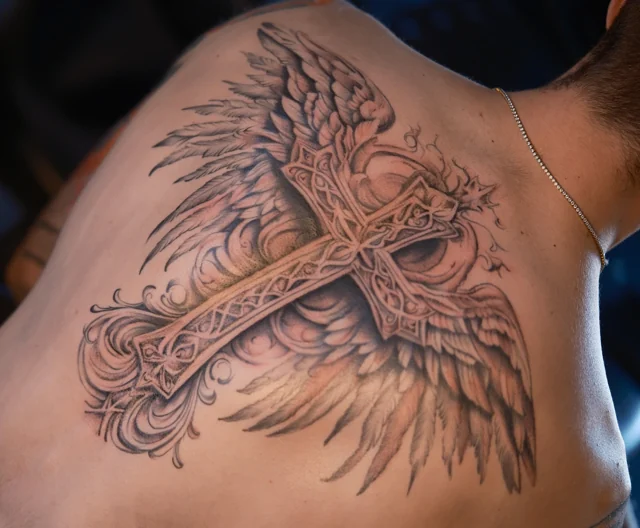 Detailed black and grey cross and wings tattoo on a shoulder